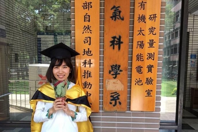 Tonia, a Taiwanese woman in a graduation gown, holding flowers and standing in front of a placard. 